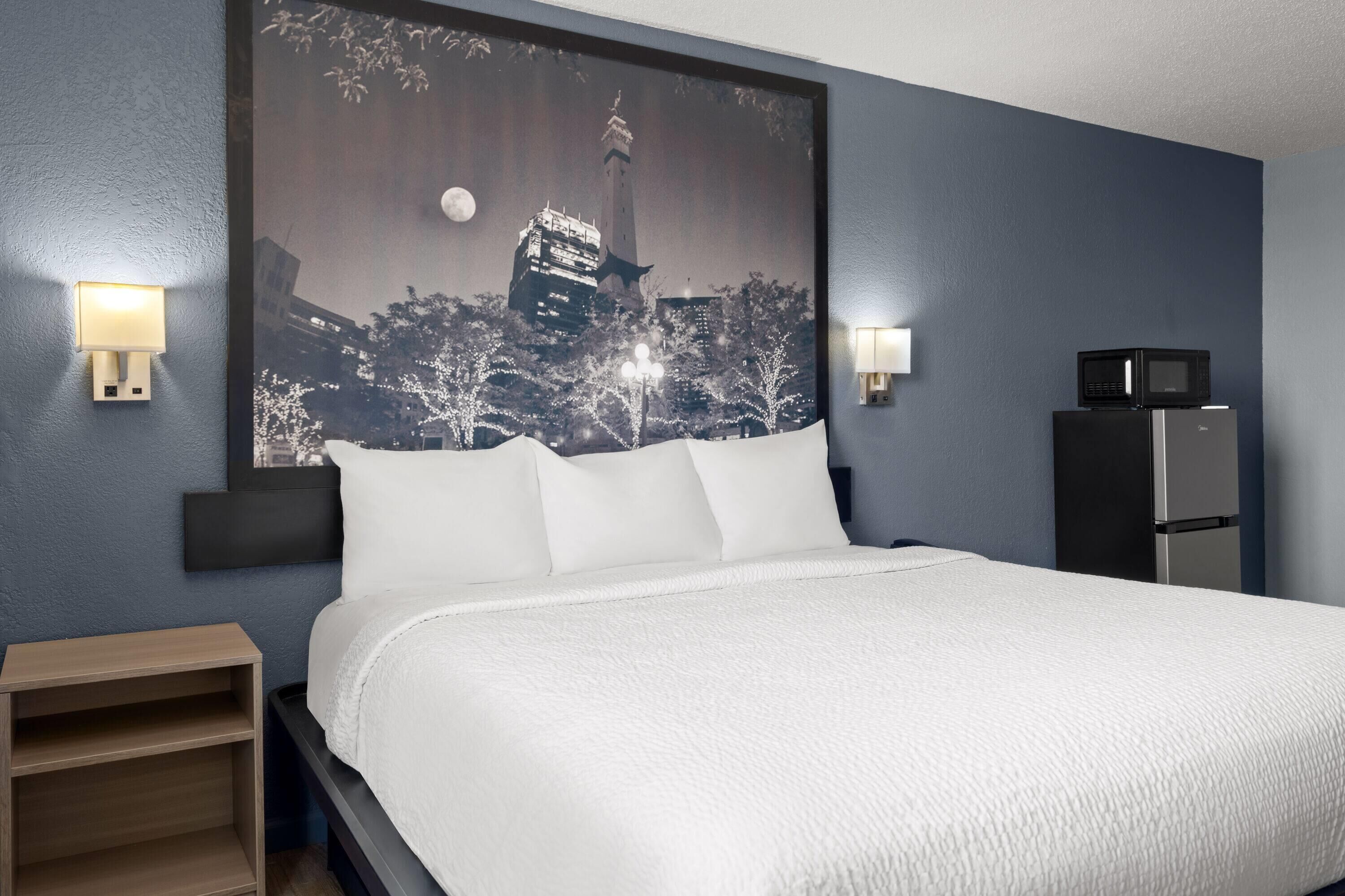 Een hotelkamer met een groot bed, twee bedlampen, een nachtkastje, een koelkast en een stadsgezicht schilderij aan de muur.