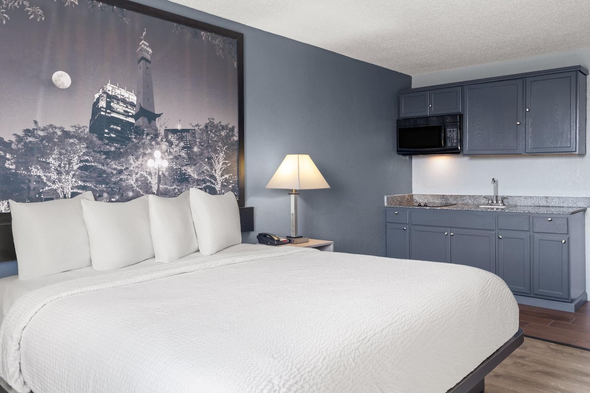 A hotel room with a large bed, a nightstand with a lamp, a microwave, and a sink in the kitchenette.