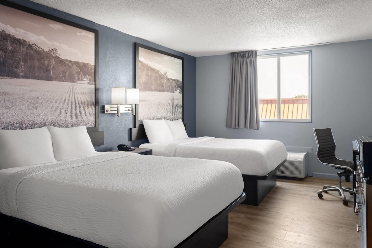 A hotel room with two beds, a desk, a chair, a large window, and two wall-mounted lamps.