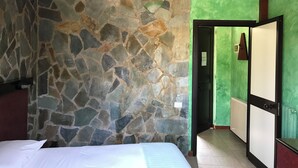 A room with a stone wall, a bed, a green wall, a door, and a mirror. - American Motel (Cisterna di Latina)