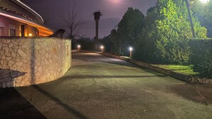 A nighttime scene of a paved pathway leading to a building with stone walls and a curved roof. Street lamps line the path, and a palm tree is visible in the background. - American Motel (Cisterna di Latina)