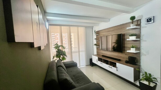 A modern living room with a dark-colored sofa, a flat-screen TV mounted on a wooden wall, and a sliding glass door leading to a balcony. - Acogedor Apto en Bello, cerca del Metro y Centro Comercial (Bello)