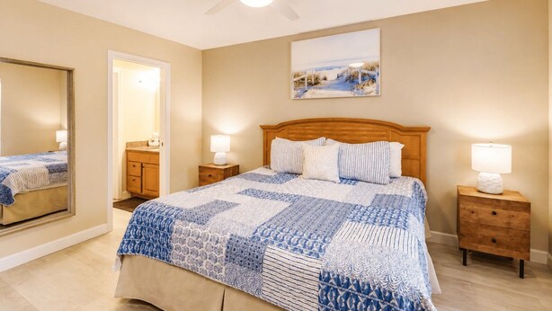 Une chambre à coucher avec un lit, une table de chevet, un miroir, un ventilateur de plafond et un tableau accroché au mur.