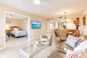 A spacious living area with a sofa, armchair, and glass coffee table. A dining area with wicker chairs and a table is visible. A bedroom with a bed and a painting is also seen. - Park Shore Resort, BLDG G #241*Great Views* (Park Shore Resort)