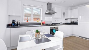 A modern kitchen with white cabinetry, a central island with seating, and a window with blinds. - Bridge House by Tŷ SA (Newport)