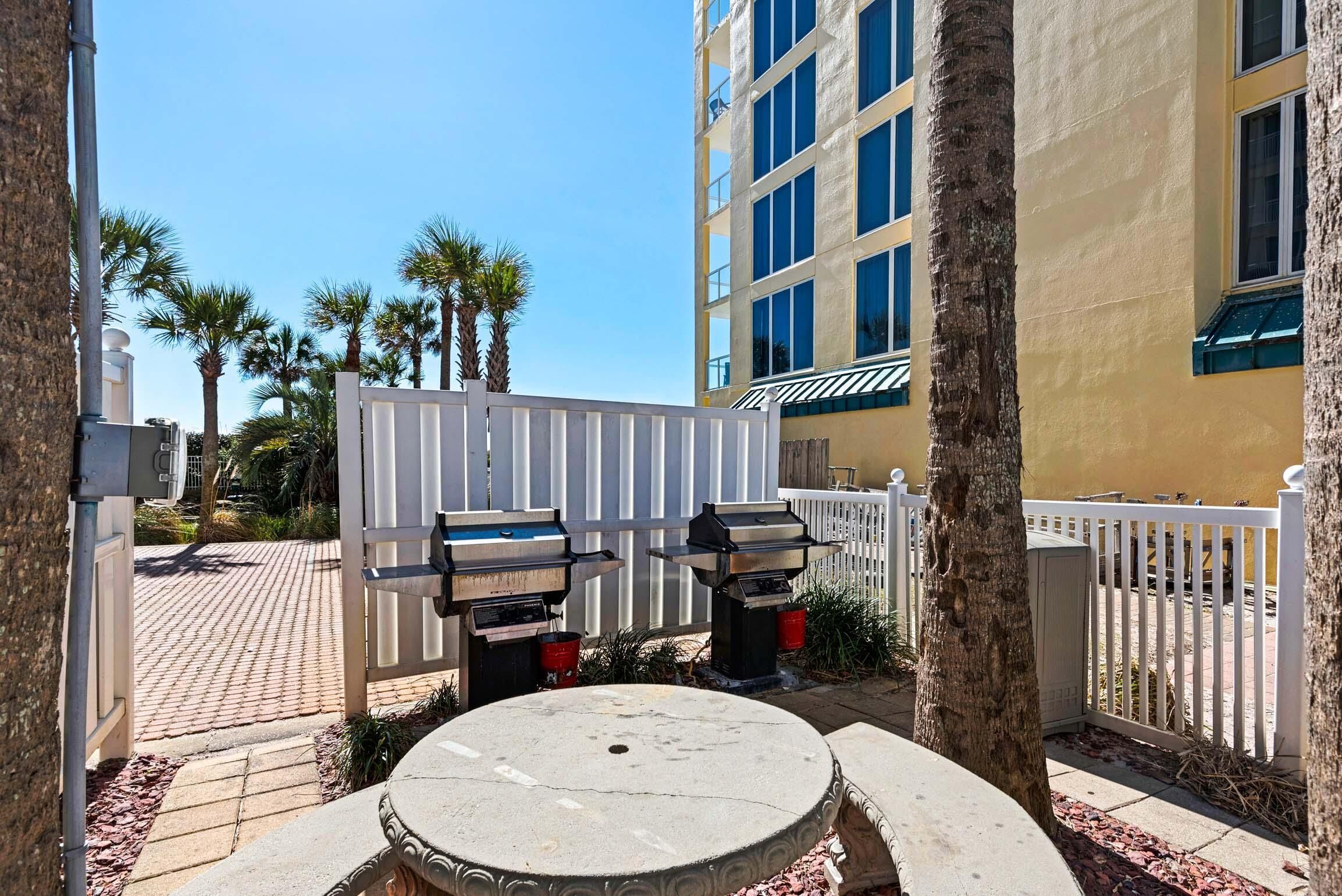 A courtyard with a white fence, two barbecue grills, a round table, and a tall building in the background.