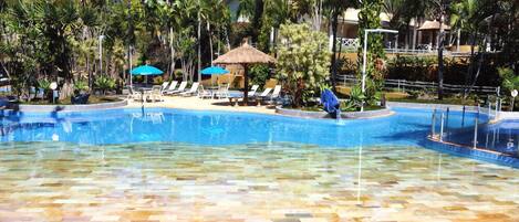 Una piscina con agua cristalina, rodeada de sillones de descanso y sombrillas, con palmeras al fondo.