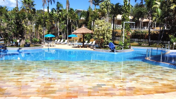 A swimming pool with clear water, surrounded by lounge chairs and umbrellas, and palm trees in the background. - Hotel Mil Flores (Braganca Paulista)