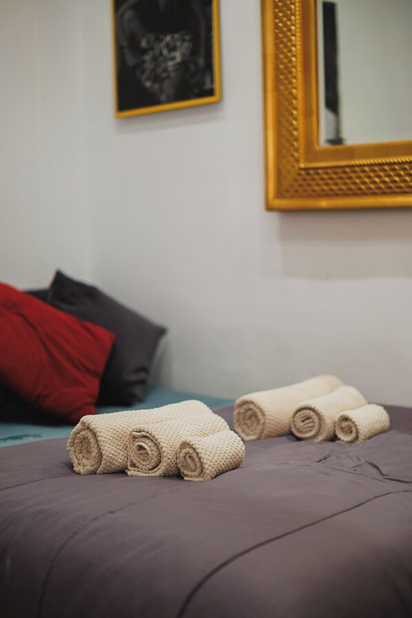 A bed with rolled towels, a red pillow, and a dark-colored blanket.
