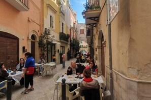 A narrow street with outdoor seating at cafes, surrounded by buildings with balconies and arched doorways.