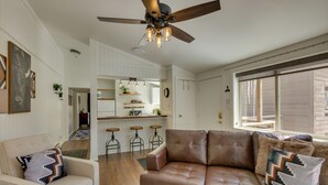 A modern living room with a brown leather sofa, a white armchair, and a patterned rug. There is a ceiling fan, a kitchen area in the background, and a large window providing natural light. - Cozy La Veta Condo: Walk to Cuchara Mountain Park! (La Veta)