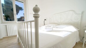 A white bed with a decorative headboard, a window with a view of buildings, and a white radiator. - Verdemare  (Chiavari)