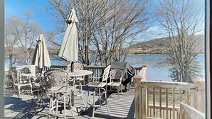 Terraza exterior con sillas y mesas de metal, una sombrilla y vista a un lago y árboles.