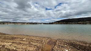 Un lago con orilla rocosa y una vista distante de casas y colinas bajo un cielo nublado.