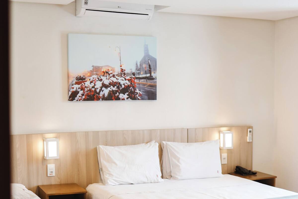 A hotel room with two beds, a wooden headboard, a wall-mounted air conditioner, a nightstand with a phone, and a painting above the beds.