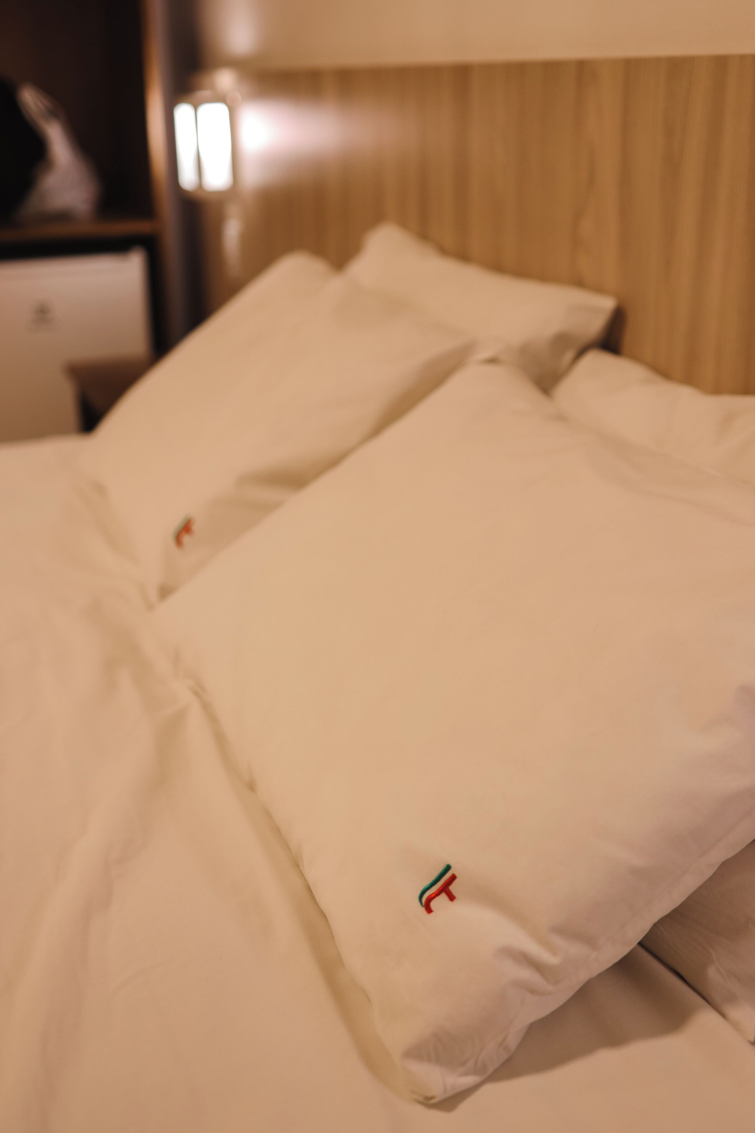 A bed with white bedding and a small embroidered logo on the pillow.