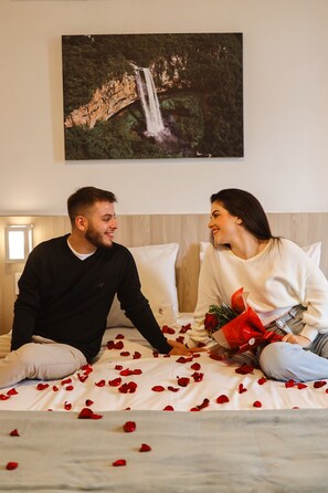 A couple sitting on a bed with rose petals, a painting of a waterfall, and a wooden headboard. - Hotel Triveneto Gramado (Gramado)