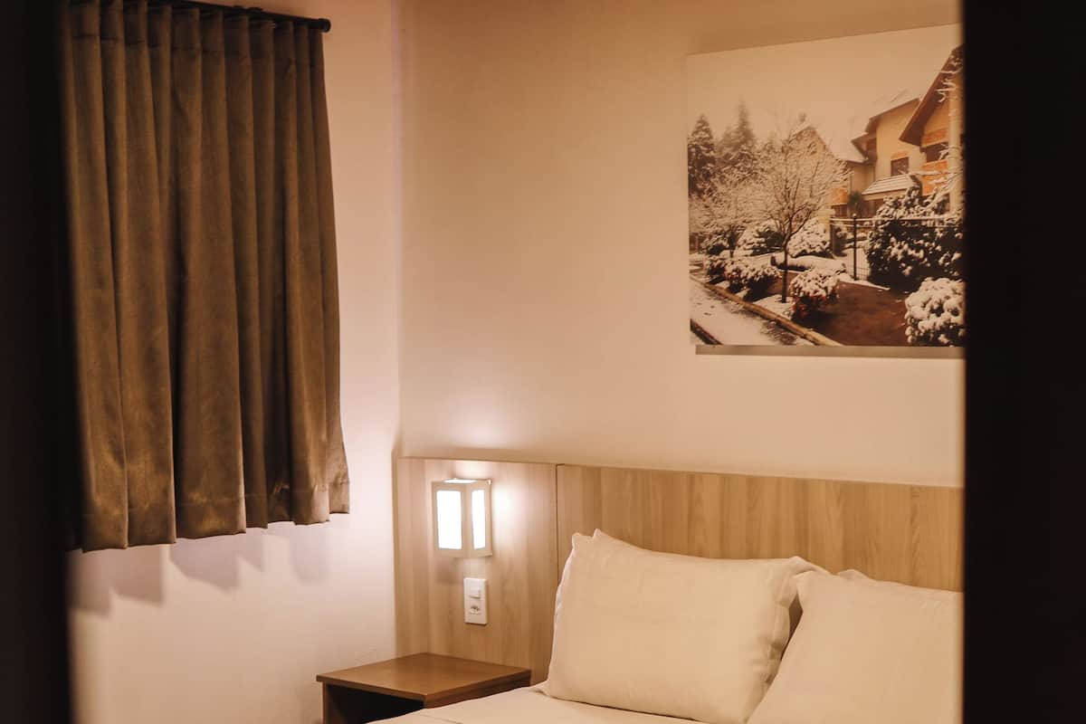 A bedroom with a bed, a nightstand, a wall-mounted light, a window with curtains, and a picture on the wall.