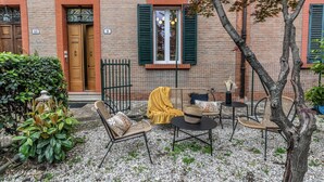 Un patio con mobiliario exterior, un edificio de ladrillo con puertas numeradas y un árbol.