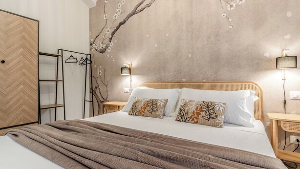 Una cama bien hecha con una manta marrón, un cabecero de madera y dos almohadas con diseños de hojas. Se observa una estantería de pared con ropa y una escalera, y una pared con un mural de una rama de árbol y flores.