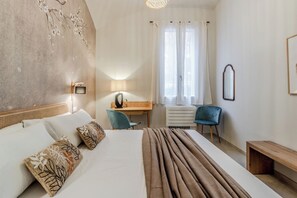 Camera d'albergo con un letto, una scrivania, una sedia, una panca e un'ampia finestra con tende.