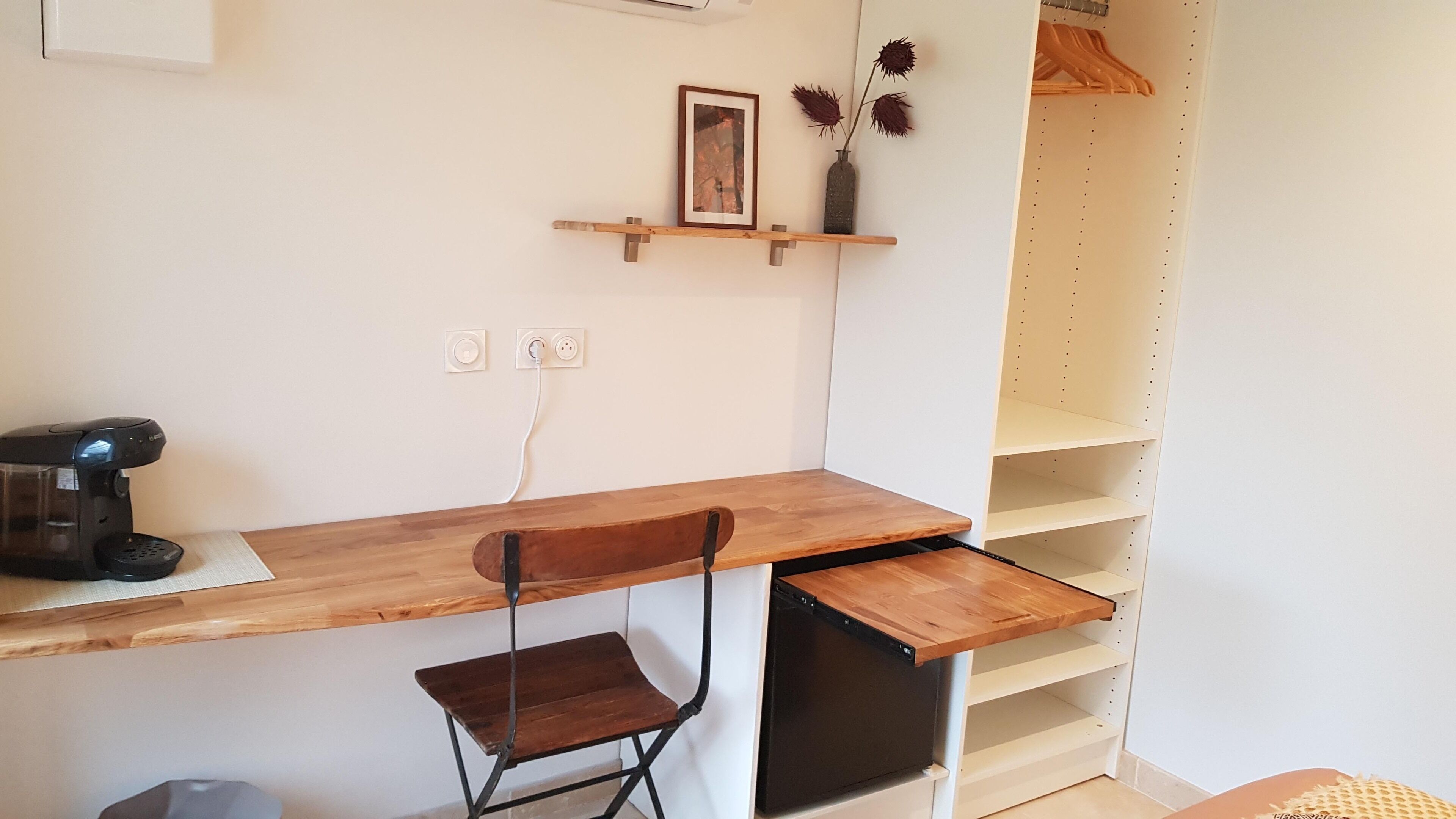 Et lille arbejdsområde med et træbord, en sort stol, en kaffemaskine og en hylderække med et billede og afdriedede planter.