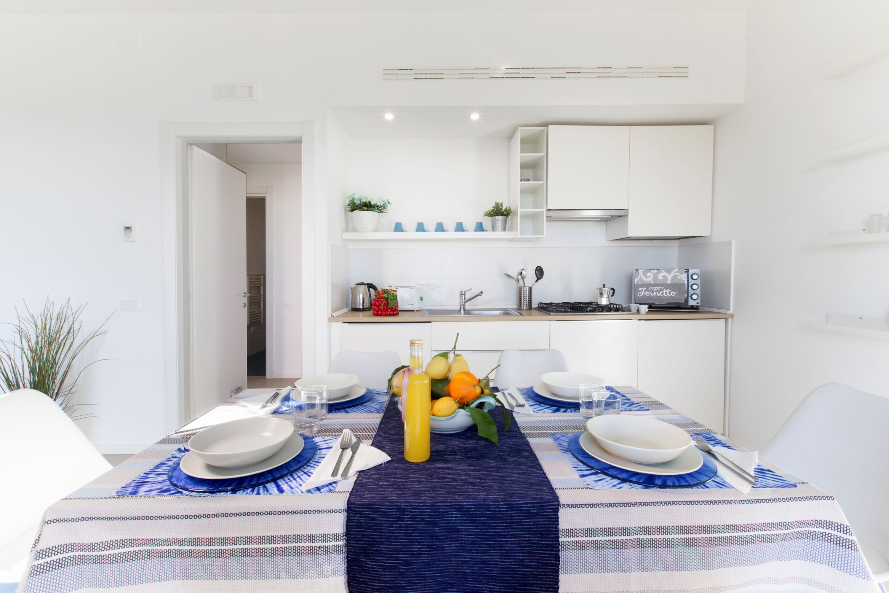 Un coin repas avec une table dressée pour quatre personnes, une cuisine en arrière-plan et une porte-fenêtre donnant sur une autre pièce.