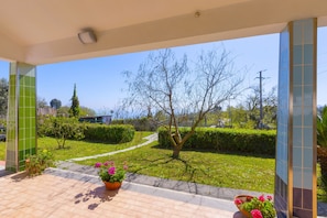 A covered outdoor area with tiled flooring, potted plants, and a view of a grassy area with trees and a pathway. - Estate4home - Villa Corinna (Massa Lubrense)