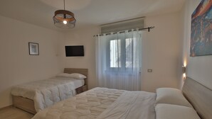 Ein Schlafzimmer mit zwei Betten, einer Deckenleuchte, einem Fenster mit Vorhängen und einem an der Wand befestigten Fernseher.