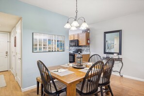 Un coin repas avec une table en bois, quatre chaises, un luminaire suspendu et une cuisine en arrière-plan.