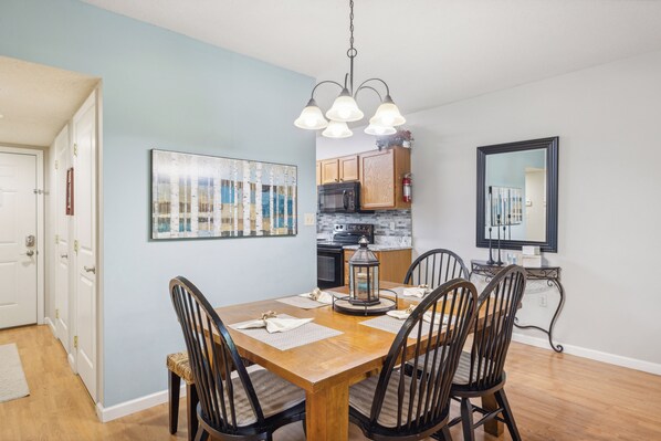 A dining area with a wooden table, four chairs, a hanging light fixture, and a kitchen in the background.