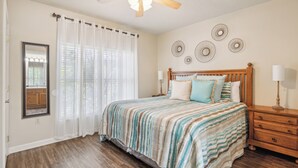 A bedroom with a bed, a nightstand, a mirror, a ceiling fan, and a window with sheer curtains.