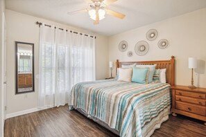 Une chambre à coucher comprenant un lit, une table de chevet, un miroir, un ventilateur de plafond et une fenêtre avec des rideaux légers.