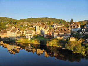 A riverside village with stone houses, a calm river reflecting the buildings, and a hilly backdrop.