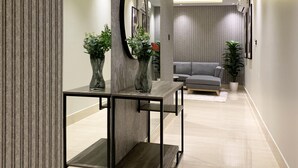 Un couloir moderne avec un canapé d’angle gris, un miroir rond et deux vases ornés de plantes.