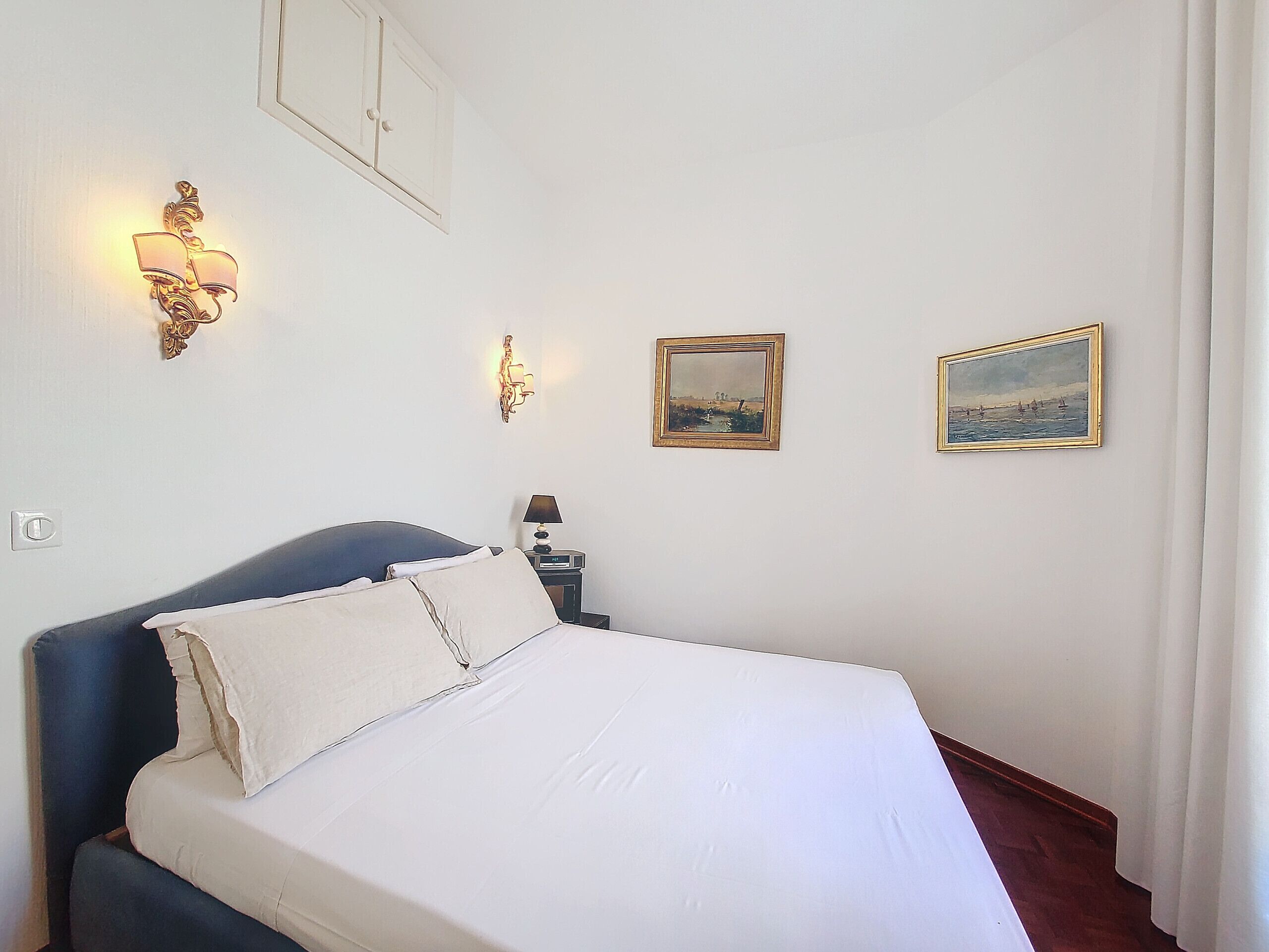 A neatly made bed with white linens, a dark blue headboard, and two wall-mounted light fixtures.