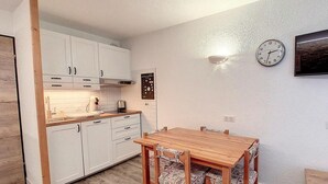 A compact kitchen with white cabinets, a wooden dining table with patterned chairs, and a wall clock. - 200m from ski slopes, 1st floor, balcony, tv, ski locker, 26m², La Rosière (La Rosière Montvalezan)