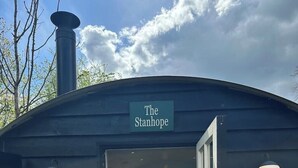 A wooden structure labeled "The Stanhope" with a door open, revealing a kitchen area with stools and a countertop. - Remote hut set in the beautiful Kent Downs (Sittingbourne)