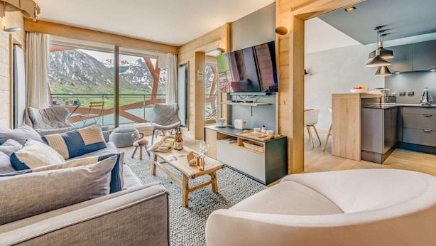 A modern hotel room with a large window offering a mountain view, a cozy seating area, a small kitchen, and a flat-screen TV.