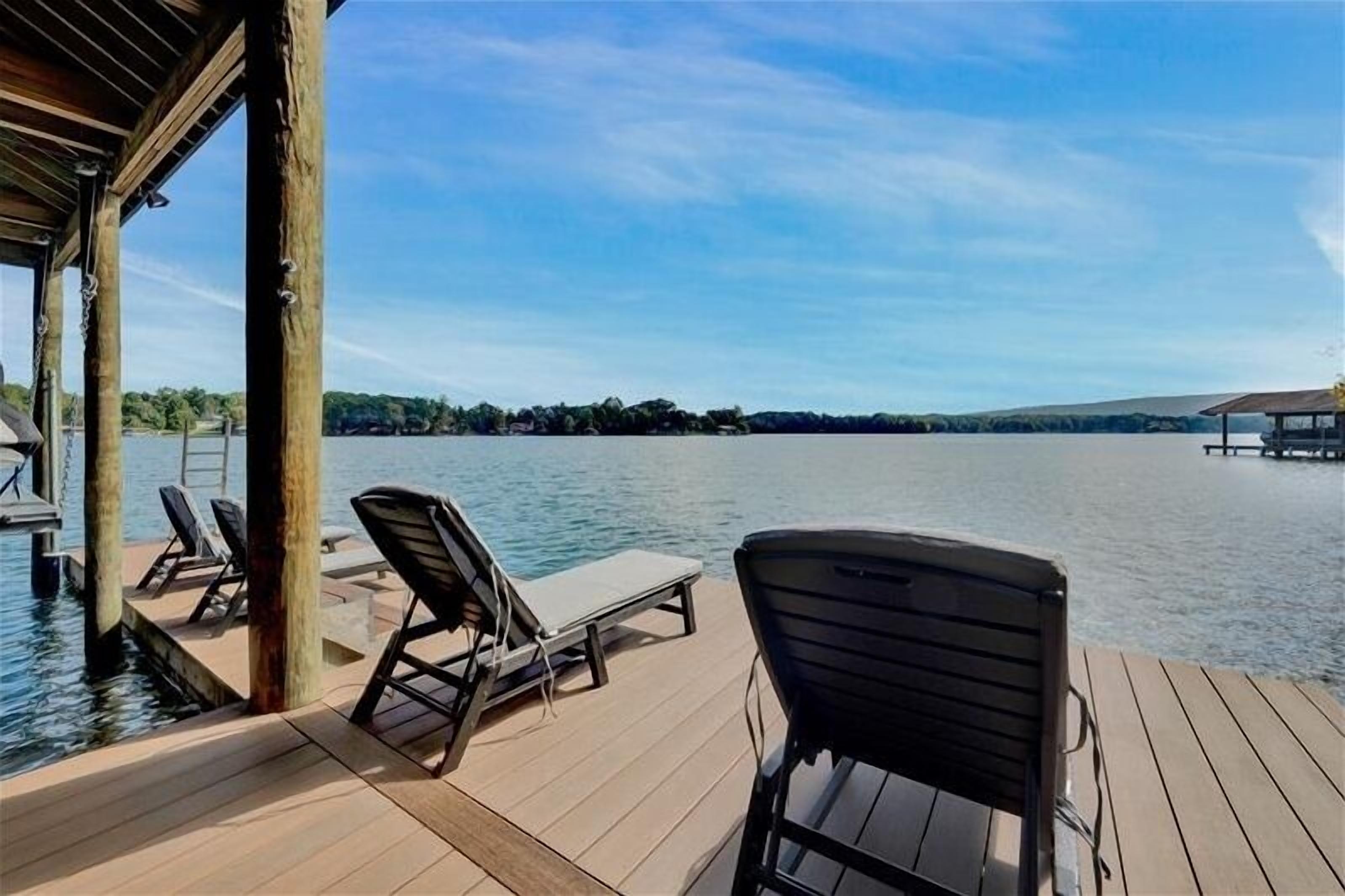 A wooden deck with lounge chairs overlooking a calm lake, with a dock and a covered area visible in the background.