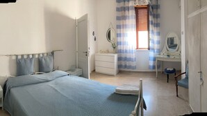 Een slaapkamer met een bed, nachtkastje, kapstok, spiegel en raam met gordijnen.