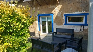 Un patio con banca y mesa, un arbusto y un edificio con detalles azules y una ventana.
