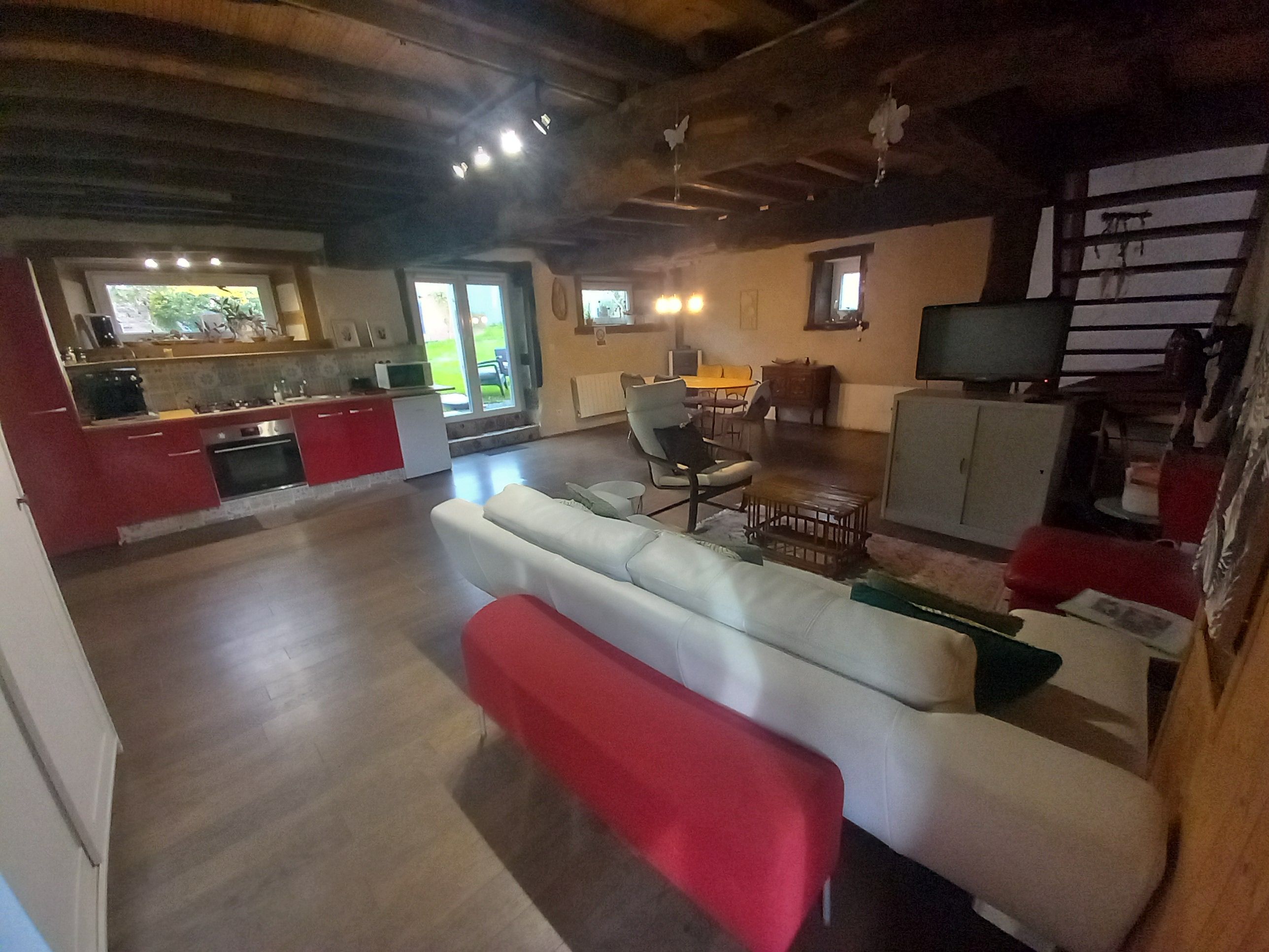 Een ruime woonkamer met een bank en een rode hocker, een keuken met rode keukenkastjes, en een eethoek met een tafel en stoelen.