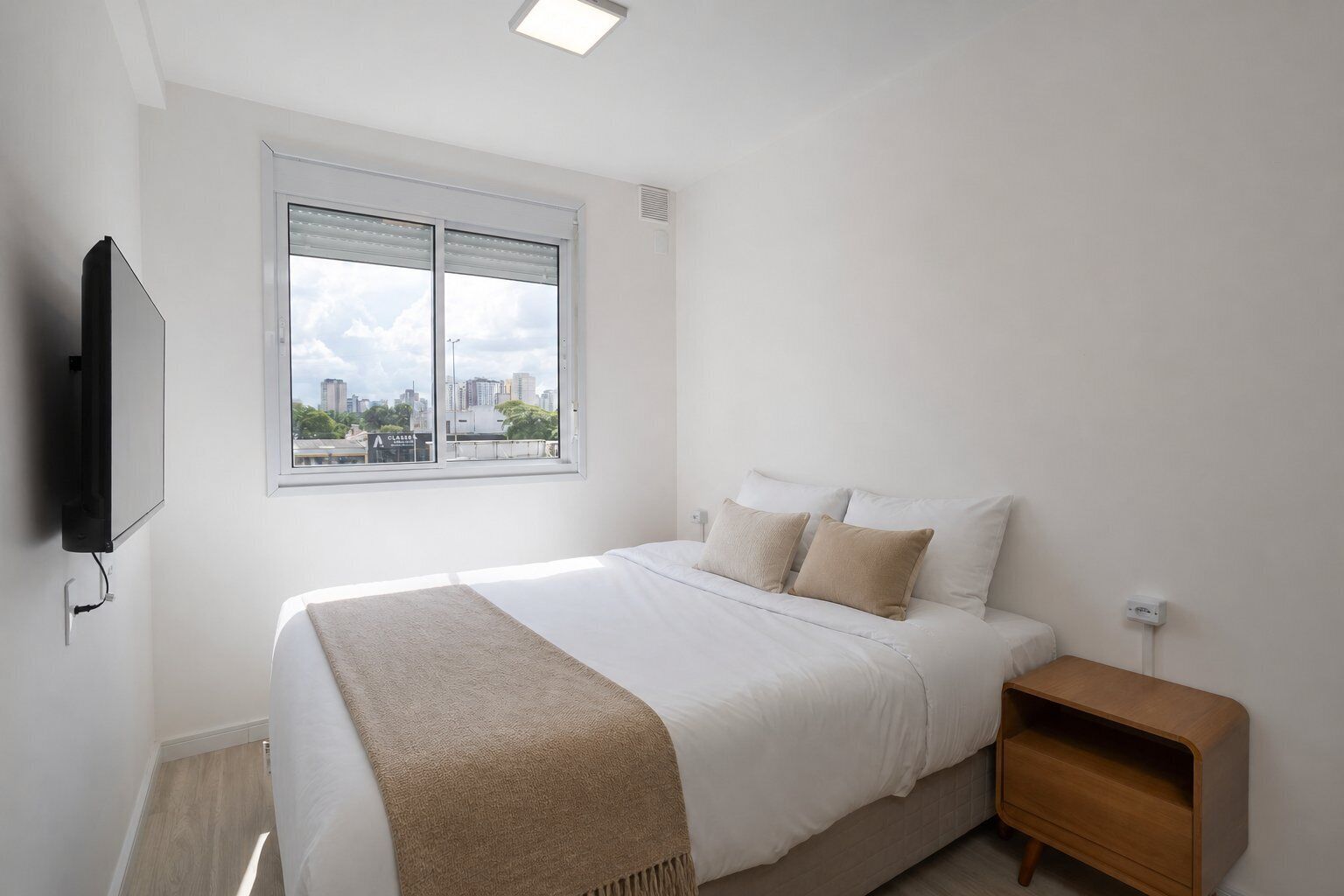 A bedroom with a bed, a nightstand, a TV, and a window with a city view.