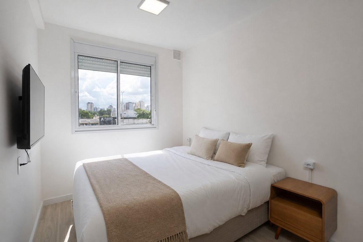A bedroom with a bed, a nightstand, a TV, and a window with a city view.