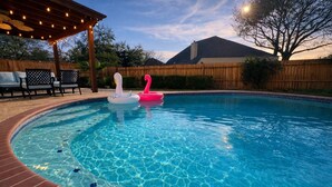 A swimming pool with two inflatable flamingos, a wooden deck, and a covered seating area with string lights. - Amazing Bay Villa Near NASA with Pool & Pool Table (Houston)