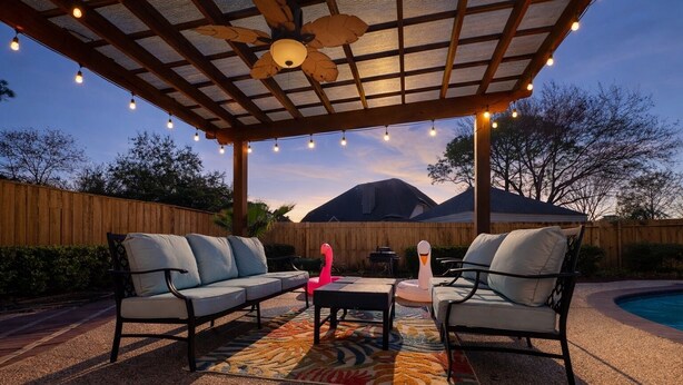 An outdoor patio with a ceiling fan, string lights, a pool, and a wooden fence. - Amazing Bay Villa Near NASA with Pool & Pool Table (Houston)