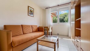 Ein Wohnzimmer mit einer Couch, einem Glastisch, einem Esstisch mit Stühlen und einem Fenster mit Vorhängen.
