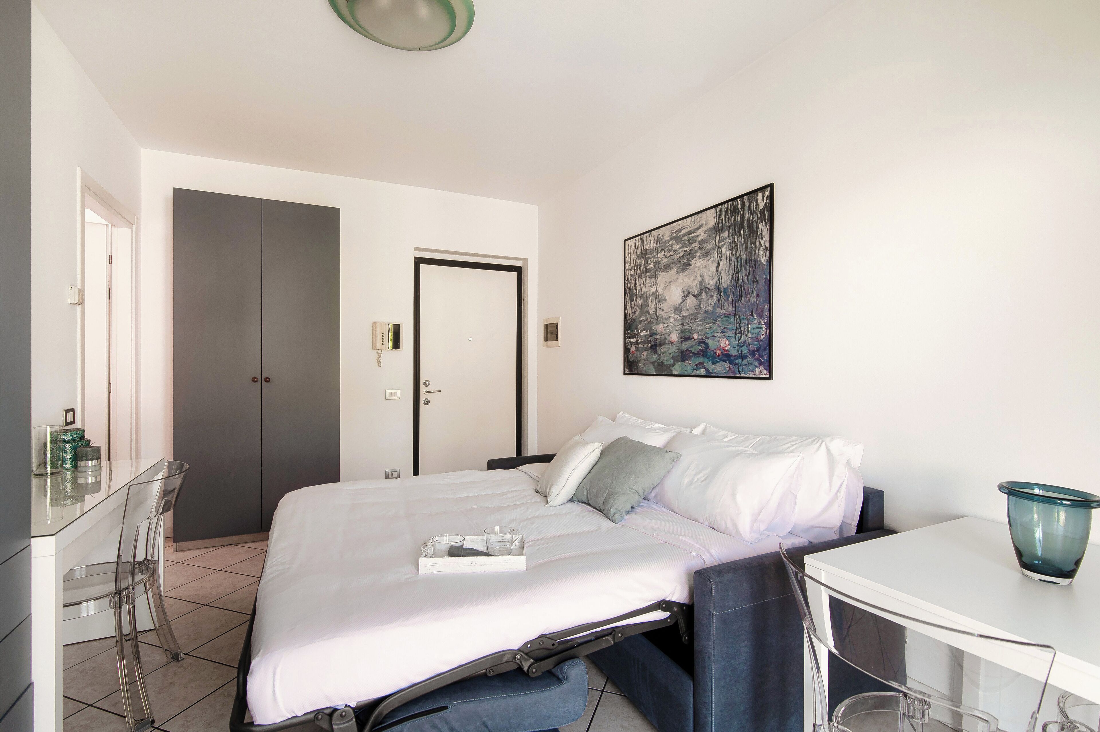 Un dormitorio moderno con una cama, un escritorio y una silla. Hay una puerta y un cuadro en la pared.