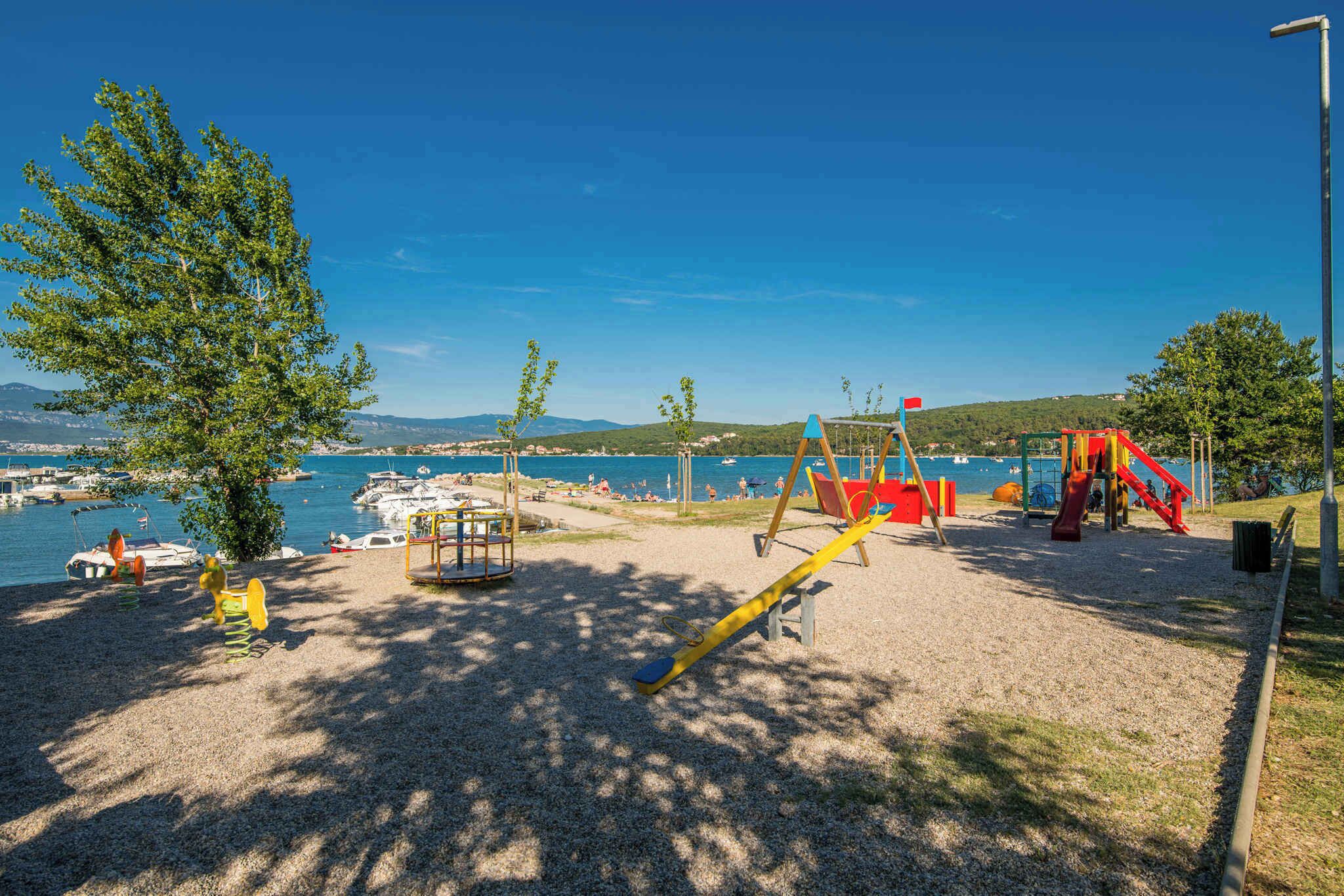 A playground with swings and slides, a tree, and a view of the water and boats.